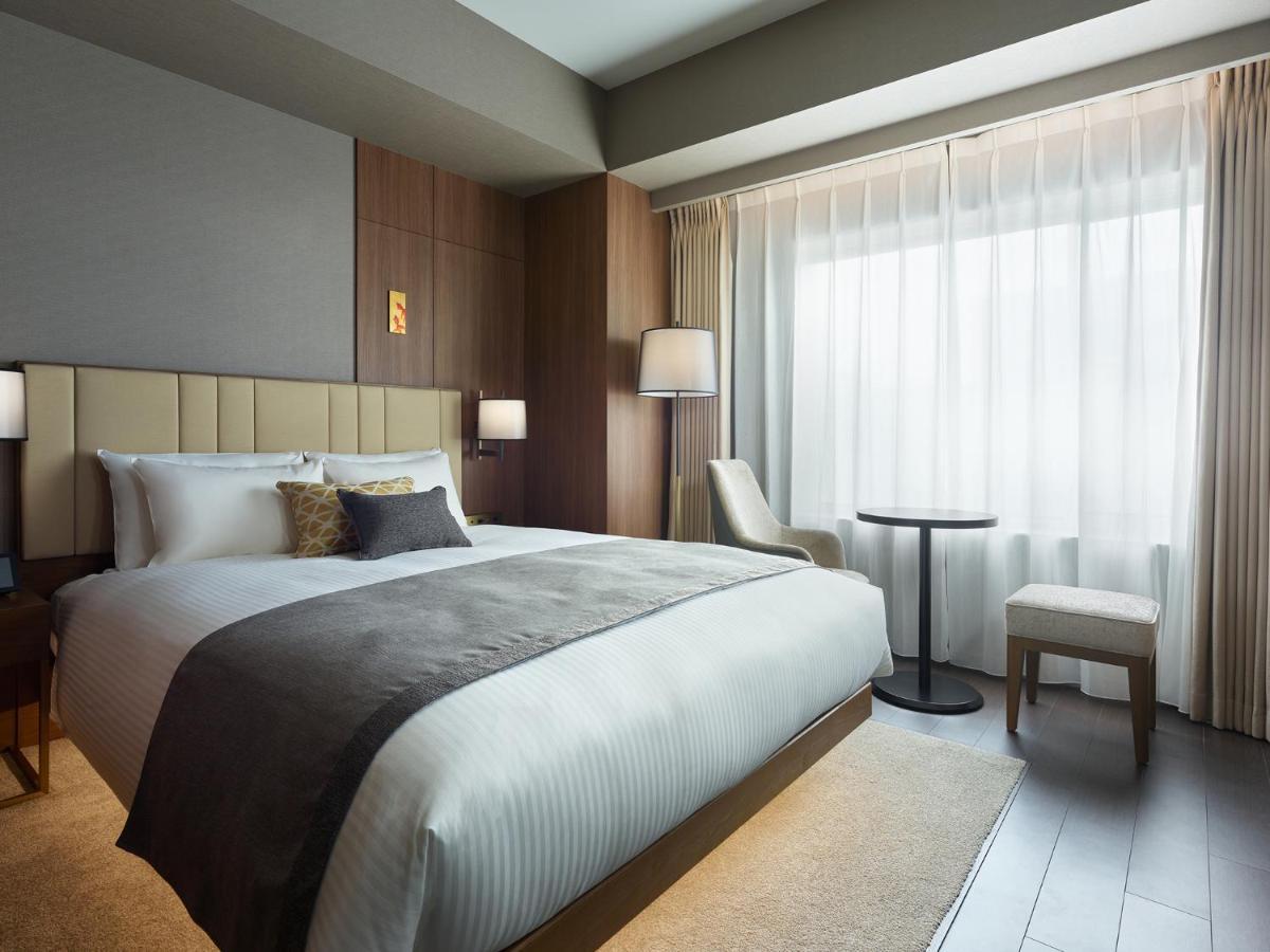 Mitsui Garden Hotel Roppongi Tokyo Premier Kültér fotó The photo shows a modern hotel room featuring a comfortable bed with white linens and a decorative throw pillow. The room has a neutral color palette with wooden accents on the walls. There are two bedside lamps and a chair positioned next to a small