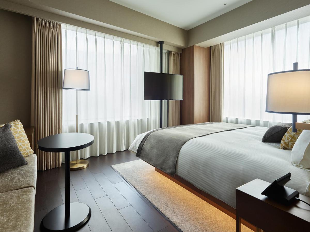 Mitsui Garden Hotel Roppongi Tokyo Premier Kültér fotó The photo shows a modern hotel room with a contemporary design. It features a large bed with crisp white linens, sitting against a wall with floor-to-ceiling windows covered by sheer curtains, allowing natural light to filter in. To the side, there's