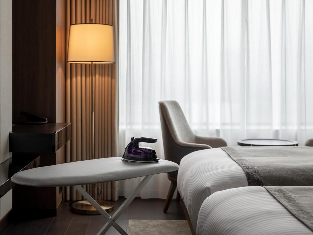 Mitsui Garden Hotel Roppongi Tokyo Premier Kültér fotó The photo shows a modern hotel room featuring two beds with white bedding. There is a light-colored armchair next to a small side table. On the table, there is an iron, and beside it, a lamp with a warm glow is illuminating the space. The large windo
