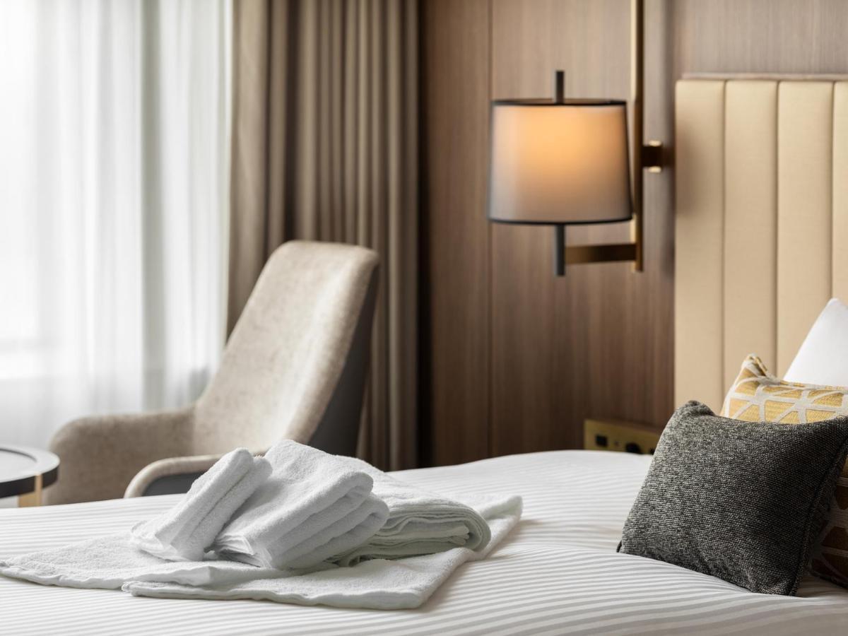 Mitsui Garden Hotel Roppongi Tokyo Premier Kültér fotó The image shows a stylish hotel room. In the foreground, there is a neatly made bed with white linens, featuring a stack of white towels arranged in a decorative manner. To the side, there is a gray upholstered armchair and a small table. The backgro