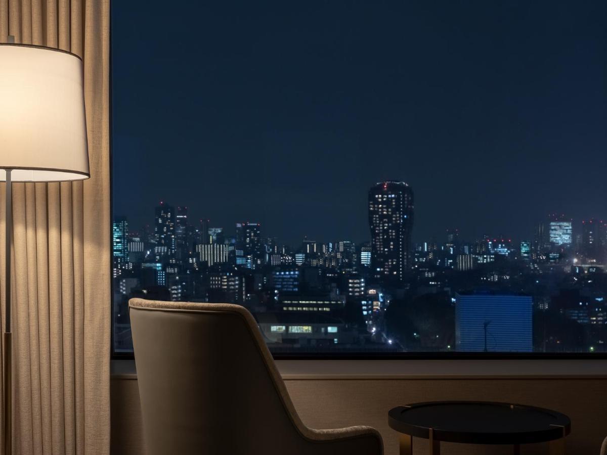 Mitsui Garden Hotel Roppongi Tokyo Premier Kültér fotó The photo depicts a cozy hotel room setting at night. In the foreground, there's a stylish armchair next to a small round table. A standing lamp provides soft lighting, creating a warm ambiance. In the background, large windows reveal a city skyline 