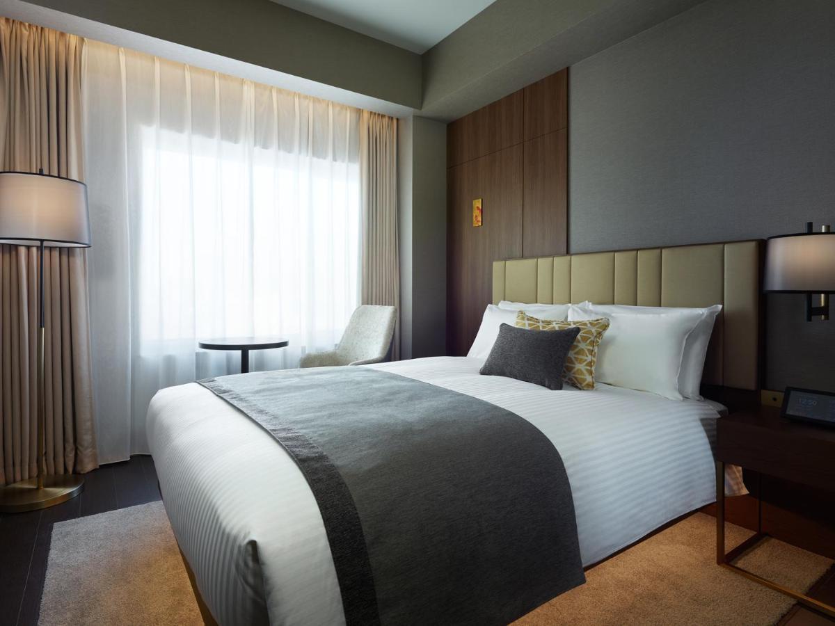Mitsui Garden Hotel Roppongi Tokyo Premier Kültér fotó The photo shows a modern hotel room featuring a neatly made bed with white linens and a decorative gray throw blanket. To the side, there is a small round table and a comfortable chair, creating a cozy seating area. The room is well-lit with natural 