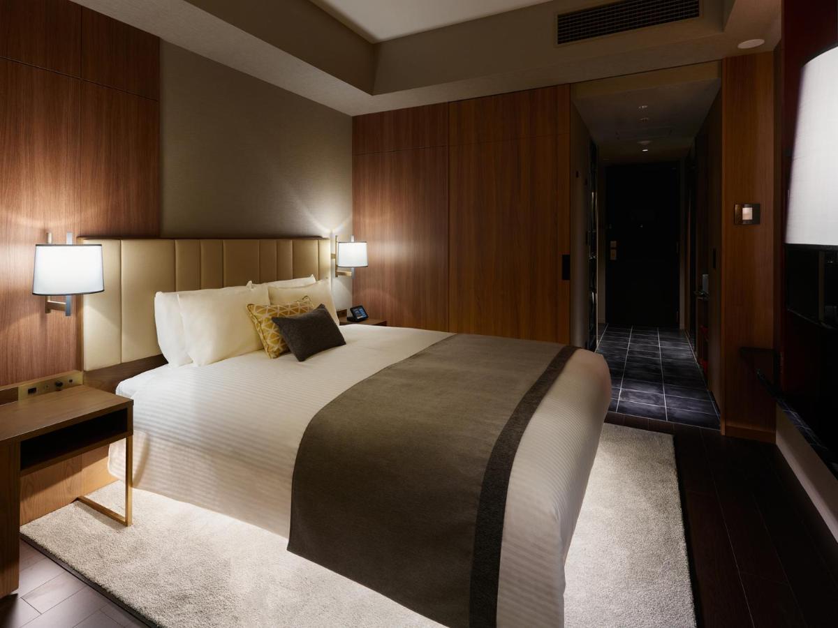Mitsui Garden Hotel Roppongi Tokyo Premier Kültér fotó The image shows a modern hotel room with a warm and elegant design. In the center, there is a neatly made bed with white linens and a dark gray throw blanket. Flanking the bed are two bedside lamps providing soft lighting. The walls have a rich wood 