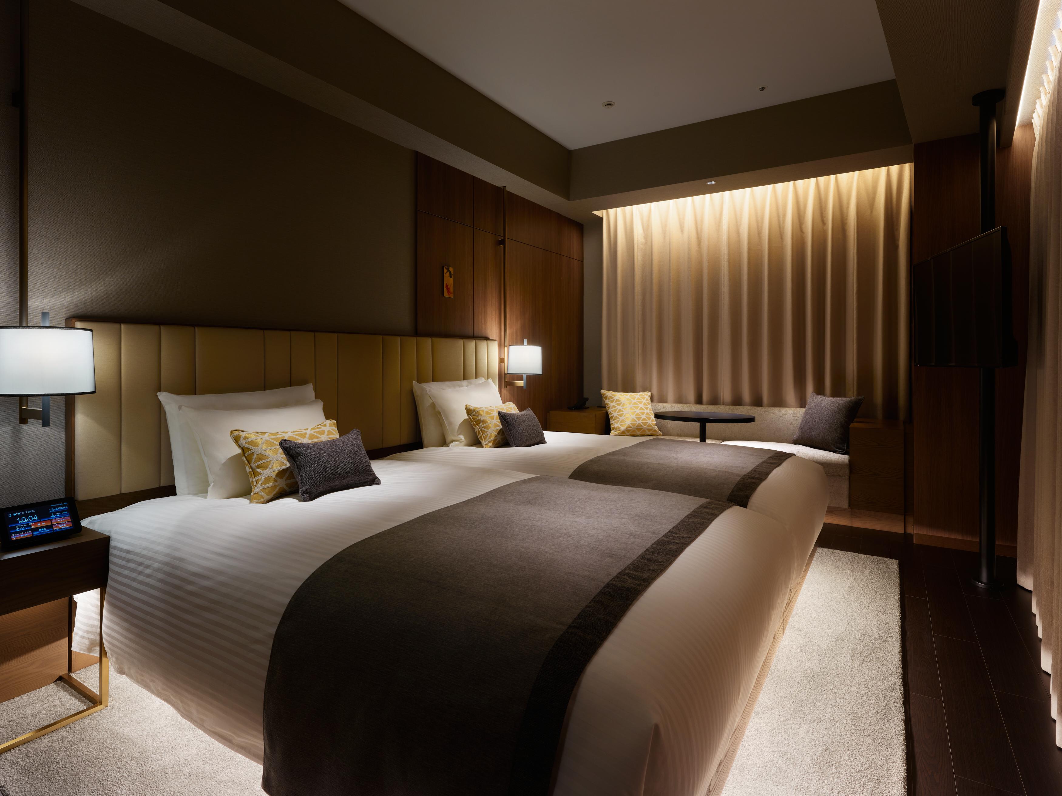 Mitsui Garden Hotel Roppongi Tokyo Premier Kültér fotó The photo shows a modern hotel room featuring two beds with plush white linens and decorative pillows. The room has a warm and inviting ambiance, highlighted by soft lighting from lamps and the backlit curtains. There is also a small table with chair
