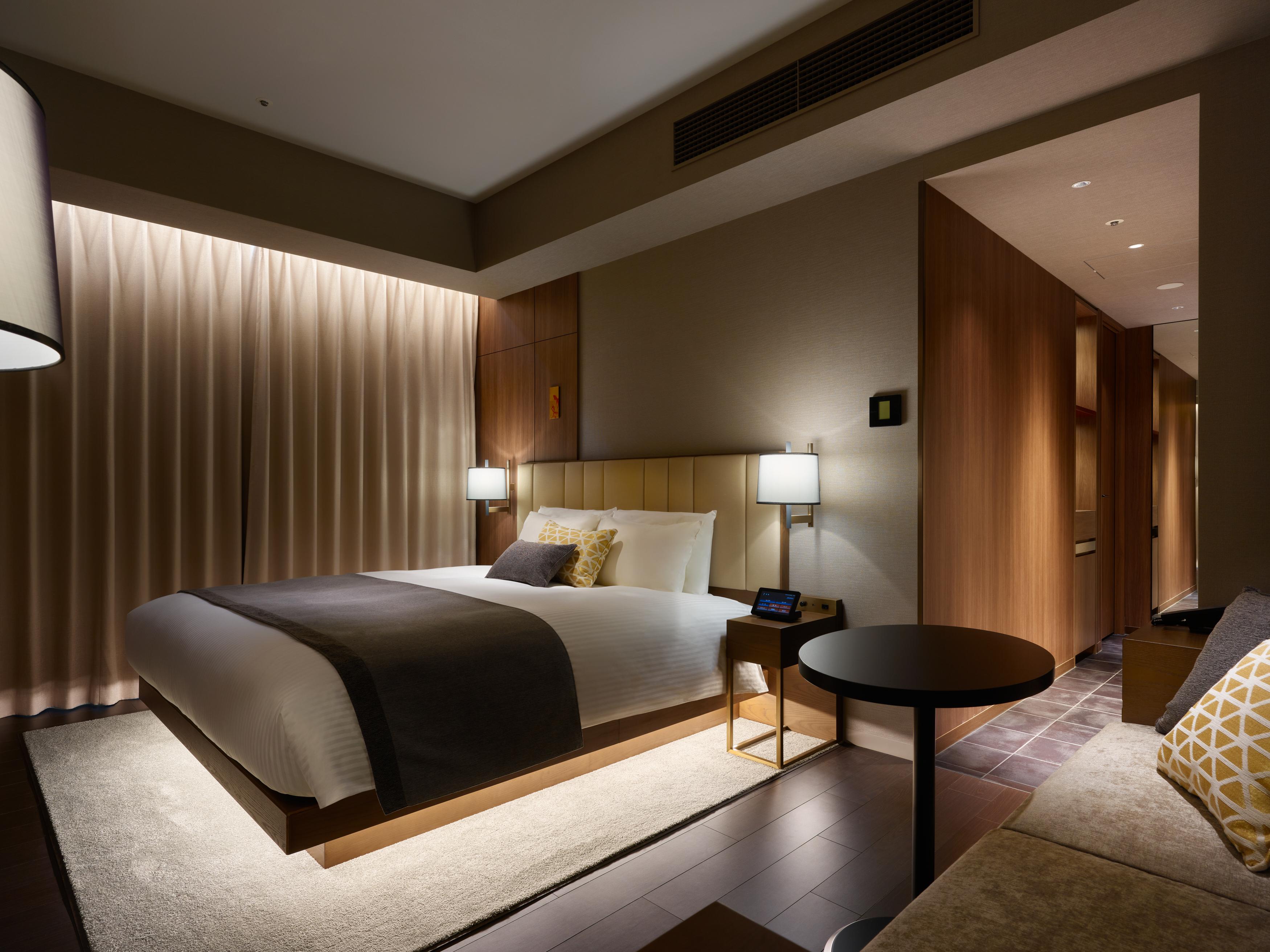 Mitsui Garden Hotel Roppongi Tokyo Premier Kültér fotó The photo shows a modern hotel room featuring a king-sized bed with neutral-colored bedding and accent pillows. There are two bedside lamps on either side of the bed, providing a warm glow. The walls have a wood finish, and there's a soft area rug on