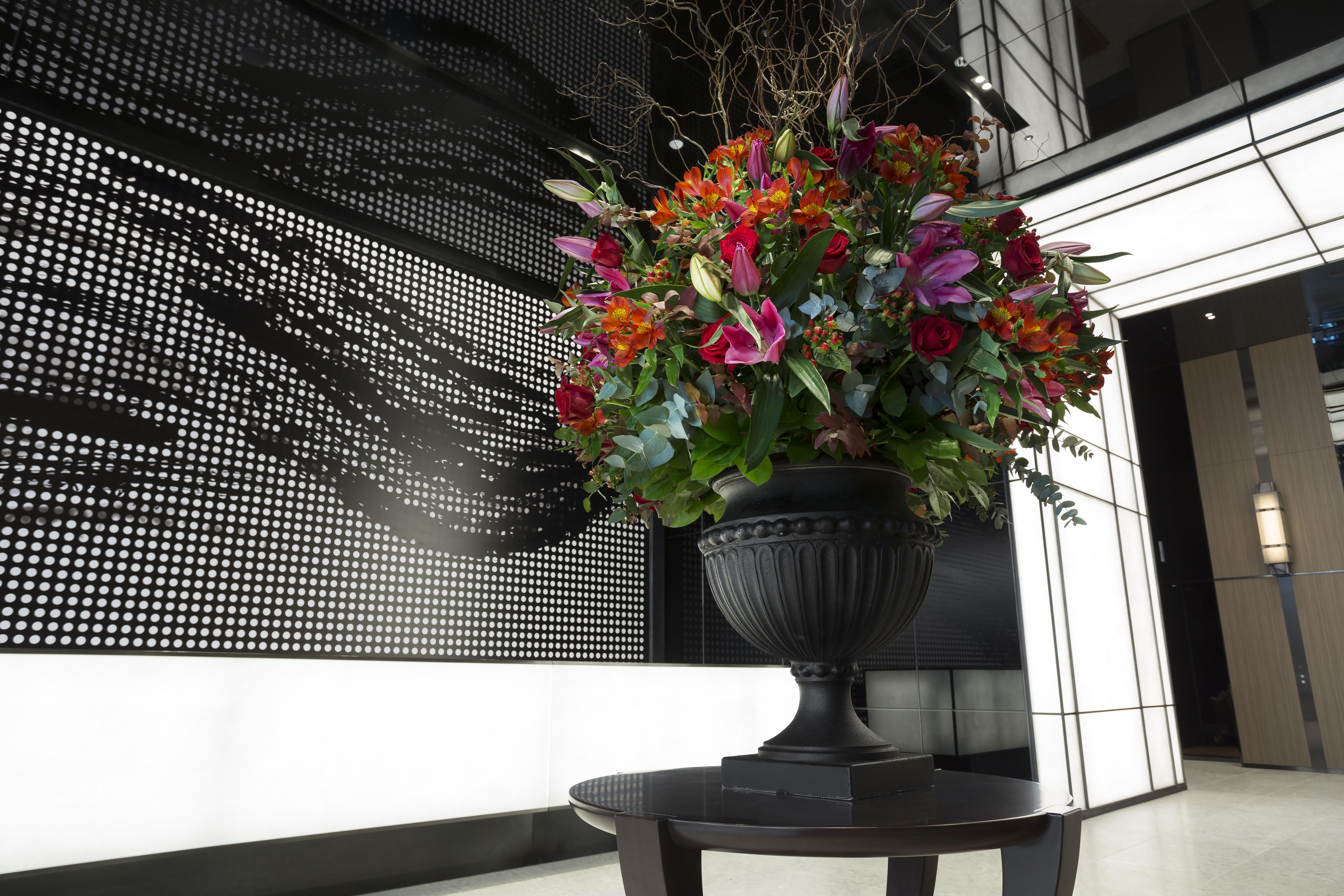 Mitsui Garden Hotel Roppongi Tokyo Premier Kültér fotó The photo features a large, vibrant floral arrangement in an ornate black urn. The arrangement is composed of various colorful flowers and greenery, showcasing shades of red, pink, orange, and green. This floral display is positioned on a small table