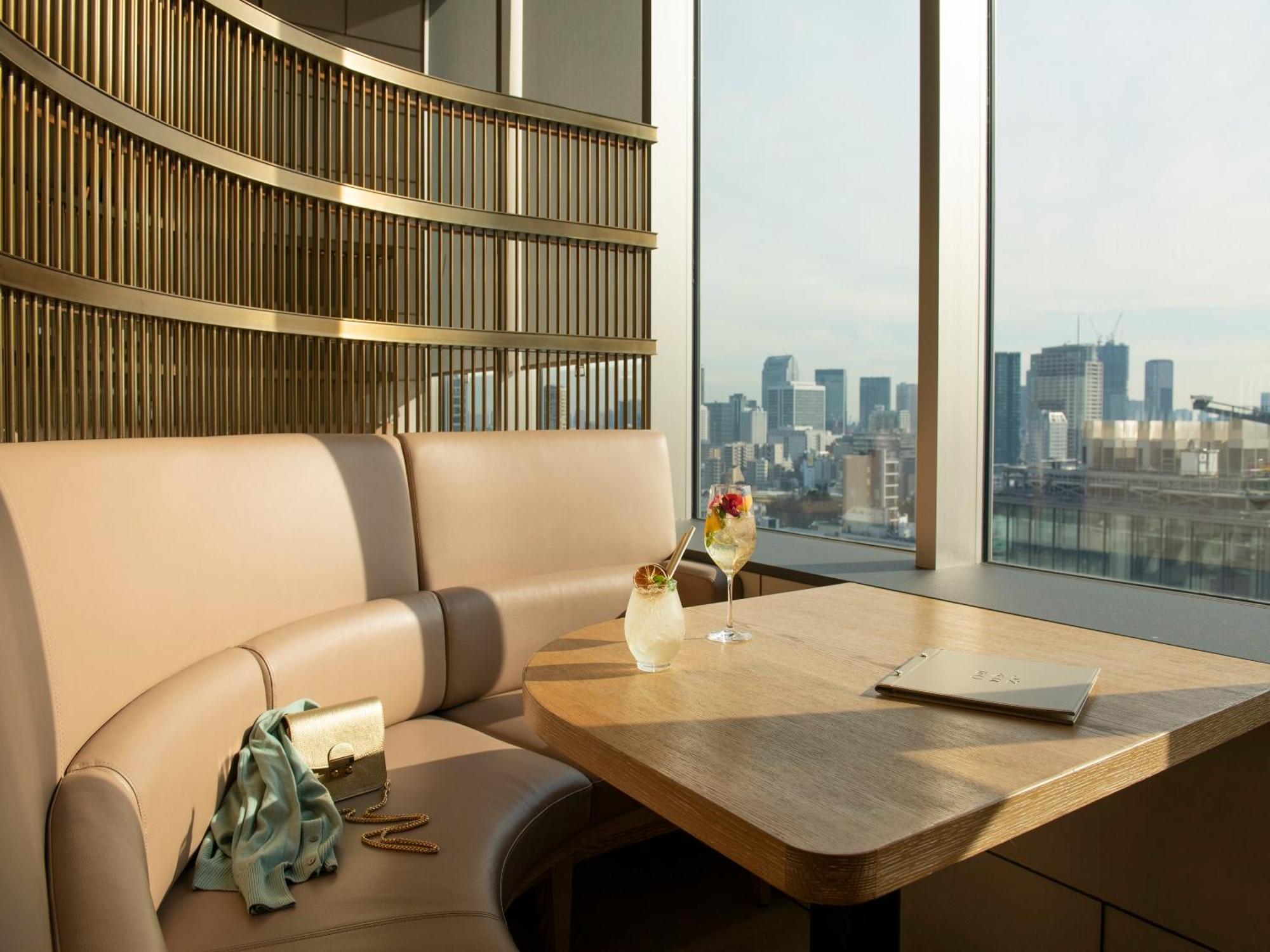 Mitsui Garden Hotel Roppongi Tokyo Premier Kültér fotó The photo depicts a cozy, modern seating area with a curved booth that features soft, pale upholstery. A wooden table is positioned in front of the booth, and on the table, there are two elegant drinks garnished with fruit. The setting has large wind