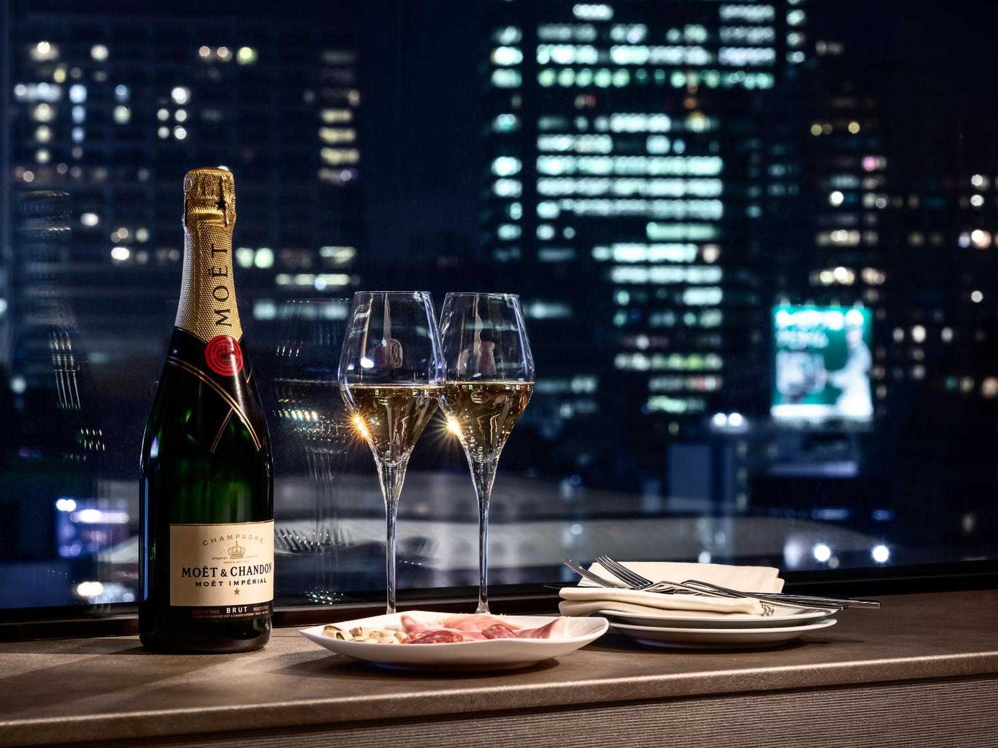 Mitsui Garden Hotel Roppongi Tokyo Premier Kültér fotó The photo features a bottle of champagne, specifically Moët & Chandon, alongside two filled champagne glasses. The table setting includes a small plate with a selection of food and neatly arranged utensils. In the background, there is a view of a cit
