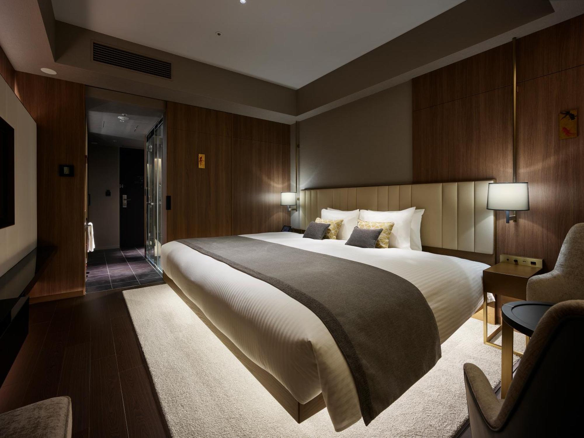 Mitsui Garden Hotel Roppongi Tokyo Premier Kültér fotó The photo shows a modern hotel room featuring a large bed with a neutral-colored duvet and a dark throw blanket. The headboard is upholstered, and there are two bedside lamps on either side of the bed. The walls have a warm wood paneling, contributin