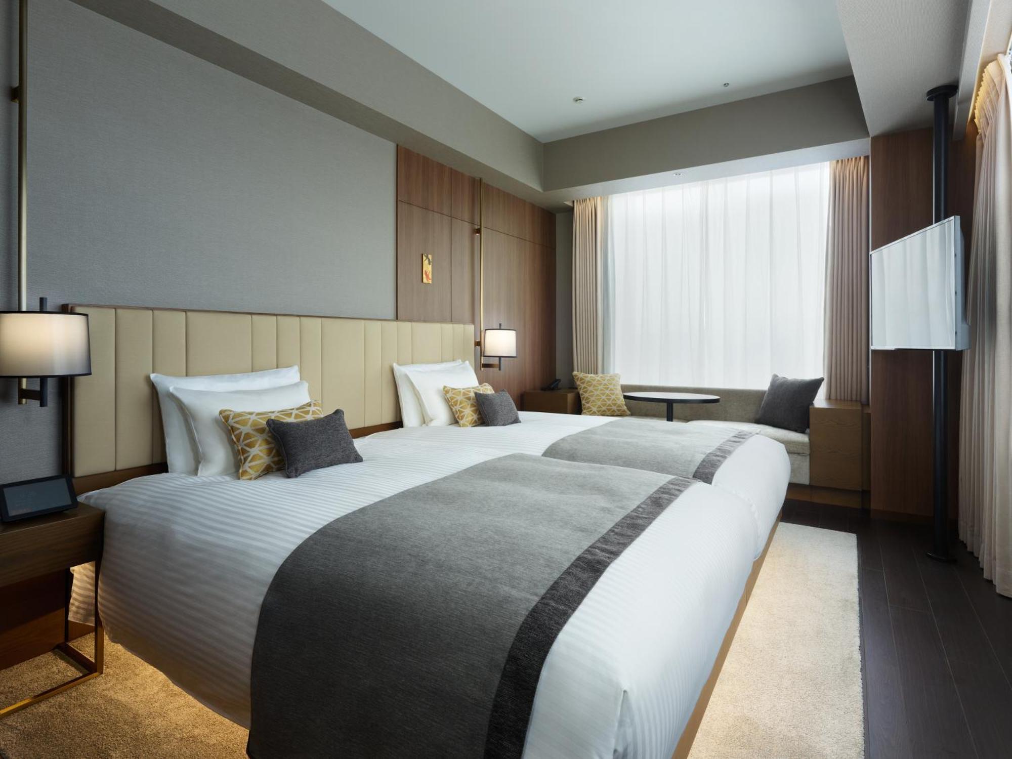 Mitsui Garden Hotel Roppongi Tokyo Premier Kültér fotó The photo shows a modern hotel room featuring two beds with white linens and decorative pillows. The room has a minimalist design, with neutral-colored walls and wooden accents. Natural light comes in through large windows, which are partially covere