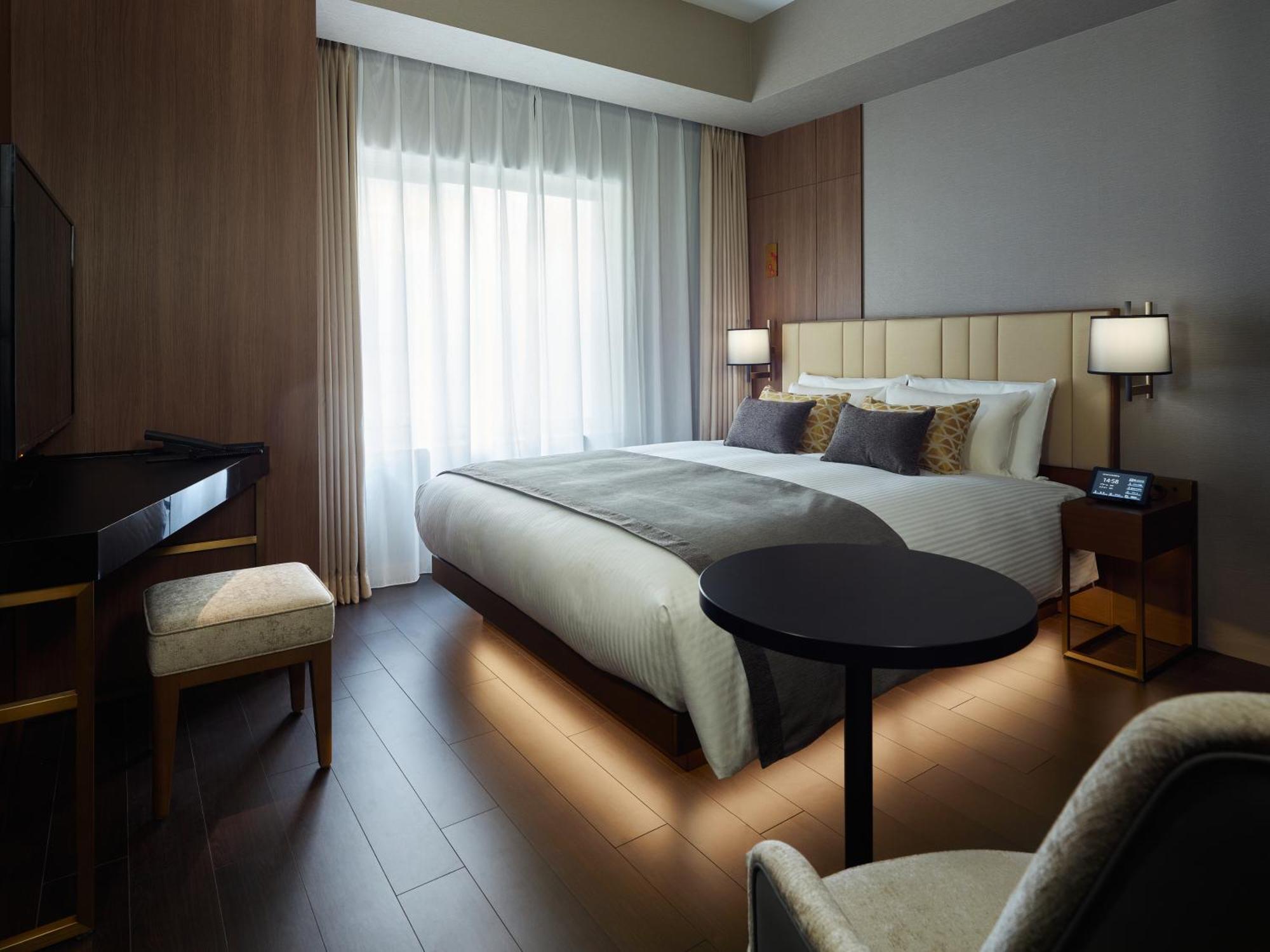 Mitsui Garden Hotel Roppongi Tokyo Premier Kültér fotó The photo shows a modern hotel room featuring a large bed with decorative pillows. There are large windows with sheer curtains allowing natural light to filter in. To one side, there is a sleek desk with a chair and a small round table in the center 